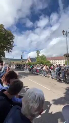 The video shows a crowd of people lining a street to watch a race of vintage cars. The cars are driving quickly down the road, and the crowd is cheering them on. The sky is blue with some white clouds. There are trees on either side of the street, and some buildings are visible in the background.
