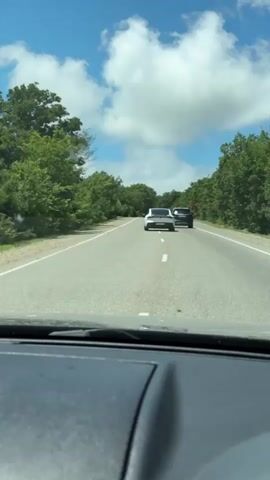 The video shows a view from the dashboard of a car traveling on a two-lane road. The road is asphalt and has a white dashed line down the center. On either side of the road are dense green trees. The sky is blue with scattered white clouds. In the distance, two cars are visible on the road ahead. The car in front appears to be white and the car behind it is dark colored. The car in the video is traveling at a moderate speed, as indicated by the movement of the scenery. The video is brief and does not contain any dialogue or sound.