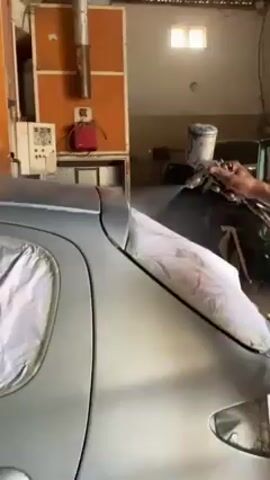 A person sprays silver paint onto the body of a silver car in a garage. The individual holds the spray gun in their right hand and moves it back and forth over the car's body, which is partially covered with paper or plastic. The environment appears to be a professional garage or workshop with various equipment, including a control panel on the wall, a ventilation duct near the ceiling, and another vehicle partially visible in the background. The light is bright inside the garage.