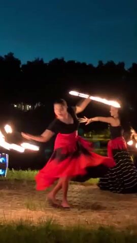 Two women are performing fire dancing outdoors at dusk. The woman in the foreground, wearing a black top and a flowing red skirt, is spinning and twirling fire poi. The woman in the background, wearing a black top and a striped black and white skirt with red accents, is also spinning fire poi. The environment appears to be a grassy area with trees and a body of water in the background. The sky is dark blue, indicating twilight. There is a speaker unit with lights visible on the left side of the frame.