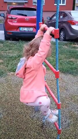A young girl with blonde curly hair is climbing a blue rope ladder with red rungs. She is wearing a pink hooded jacket and pink leggings with a strawberry pattern. Her right hand is gripping the top rung, and her left hand is reaching for the rung above. Her right leg is bent at the knee, and her left leg is extended downwards, resting on a lower rung. The rope ladder is attached to a blue pole. The ground beneath the ladder is covered in small, multicolored pebbles. In the background, a red Mazda SUV is parked. Further back, another car, possibly dark gray or brown, is visible. A building with windows is also in the background. The overall setting appears to be an outdoor playground or courtyard.