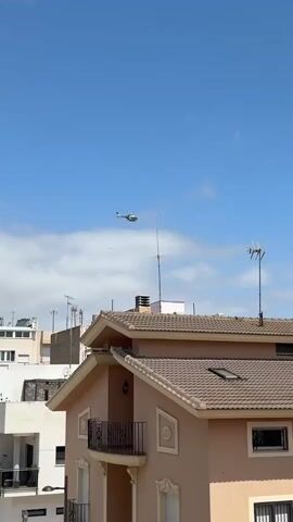 The video shows a helicopter flying across a clear blue sky with some scattered white clouds. The helicopter is moving from left to right, appearing relatively high above a residential area. In the foreground, several buildings with terracotta-tiled roofs are visible. The most prominent building has a peach-colored facade with dark railings on balconies and windows, and a few skylights on its roof. Antennas are present on the rooftops of some buildings. Palm trees are visible in the lower part of the frame, near the buildings. The scene is brightly lit, indicating daytime. No people or visible text are present. The setting appears to be a sunny residential area, consistent with the location context of Sollana, Spain.