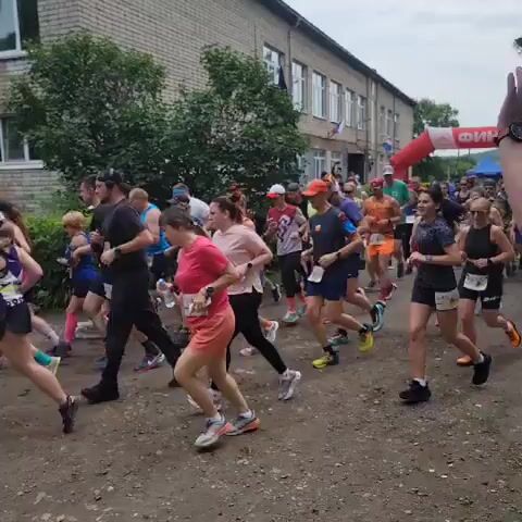 A group of people are running in a race. They are on a dirt path in front of a brick building. The people are wearing athletic clothing and some have bib numbers. The race appears to be a trail run due to the terrain. The weather seems to be overcast.