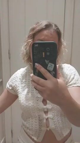 A young woman with blonde, curly hair stands in what appears to be a fitting room. She is taking a selfie with her phone. She is wearing a white, cropped, short-sleeved sweater with a button-up closure that ties with small bows, and a white mini skirt with a floral print. Her left arm is resting on a wall or doorframe, and her right arm is extended, holding the phone. To her left, on the floor, are a pair of white sneakers and a denim jacket. To her right, on the floor, is a brown handbag. The walls are a neutral, light color, and there is a wooden floor.