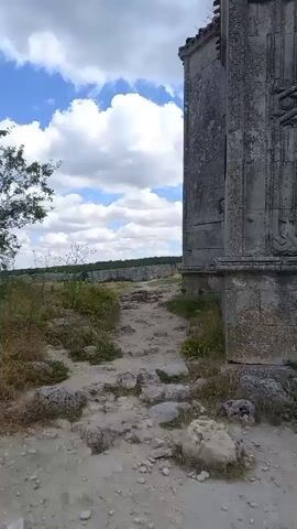 The video pans across an ancient stone structure, likely a tomb or mausoleum, with weathered carvings and a barred entrance. The structure sits on a rocky, overgrown hillside under a cloudy sky. A pathway made of stones leads up to the entrance, which is flanked by ornate pillars and decorative stonework. The overall impression is one of historical significance and natural decay.