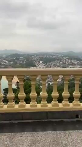 The video pans from left to right across an overlook of a beach city. A yellow balustrade is in the lower portion of the frame. Trees are to the right of the balustrade, with the buildings of the city visible past them. The bay is full of boats, and the sandy beach stretches along the arc of the bay. Mountains can be seen on the horizon. The sky is overcast.