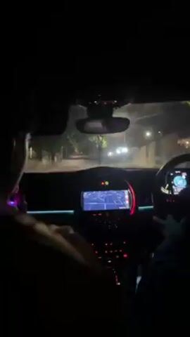 A first-person perspective shows a car driving at night on a lit street. The car's dashboard is visible with a navigation system and the steering wheel. Two people are in the car; one is driving and the other is in the passenger seat. The view looks forward out of the front windshield. Buildings and trees line the side of the road.