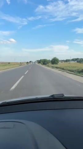 The video shows a first-person view from inside a car driving on a paved road. The road has a white dashed line separating the lanes and a solid white line on the right side. The car is moving forward. The environment outside the car appears to be rural, with fields and scattered trees on either side of the road. The sky is blue with scattered white clouds. In the distance, there are some buildings and more trees. The dashboard of the car is partially visible in the foreground. The video is shot during the daytime.