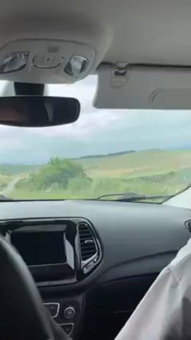 The video is taken from the interior of a car, looking out through the windshield at a rural landscape. The dashboard of the car is visible, along with the rearview mirror and sun visors. Beyond the windshield, the scene shows rolling green hills with some scattered trees under an overcast sky. The car is moving along a road, and the wipers appear to be active, suggesting light rain or mist. The driver's arm in a light-colored shirt is visible on the right side of the frame.