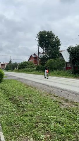 A person is riding a bicycle down a paved road. They are wearing a white top and orange shorts with a backpack. The road is lined with grass and trees. There are houses and buildings in the background. The sky is cloudy. A sidewalk is visible on the left side of the road. A person is walking on the sidewalk. The video pans to the right, showing more of the sidewalk and houses. A fence is visible in front of one of the houses, with bushes and flowers behind it. A red flag is visible on a pole behind the fence.