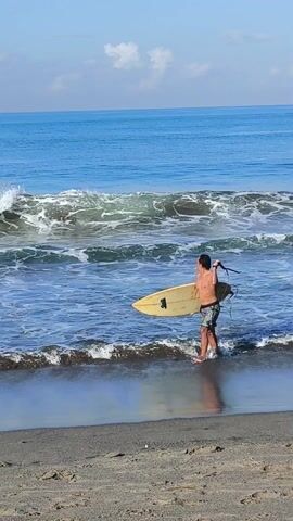 A medium shot depicts a man carrying a yellow surfboard on a beach, heading towards the ocean. The man is shirtless, wearing checkered shorts. He holds the surfboard horizontally in front of him. In the background, the ocean waves are breaking, and another person is swimming. The sand on the beach appears wet and reflective, and the sky above is clear and blue.