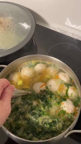 A person stirs a pot of soup with a spoon on a black stovetop. The soup contains meatballs, carrots, and green leafy vegetables. Another pot with a lid on is visible in the background. Steam is rising from the soup. The person's hand is visible while stirring the soup. The person says "Супчик бомба" which translates to "Soup is the bomb" in English.