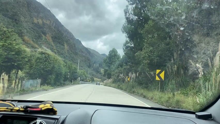 The video shows a car driving on a winding mountain road. The road is surrounded by lush green vegetation. The sky is cloudy and overcast. The car is approaching a curve in the road, marked with yellow arrow signs. There are some other cars on the road. The interior of the car is visible, including the dashboard and steering wheel. There is a black and yellow cloth on the dashboard.