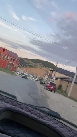 The video shows a view from inside a vehicle driving down a paved road lined with houses and buildings. The sky is partly cloudy, with hues of blue and gray suggesting it is either early morning or late evening. On the left side of the road, there are brick houses with red roofs, and on the right, there are buildings with various roof types, some appearing to be under construction or in disrepair. Several cars are visible on the road and parked along the side. The road itself is somewhat uneven. In the background, a hill with scattered houses and a telecommunications tower can be seen. The dashboard of the vehicle is partially visible in the foreground, indicating the perspective is from the driver's seat.