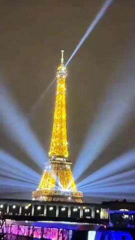 The video shows the Eiffel Tower at night, illuminated by golden lights and surrounded by blue spotlights emanating from below. The tower is a prominent structure in the center of the frame, with a bright beam of light extending upwards from its top into the dark sky. In the foreground, a dimly lit boat or ferry is visible with people on board, their faces and clothing illuminated by purple and pink lights. The overall scene is one of a nighttime spectacle, likely a tourist attraction. The audio consists of spoken words in Turkish, discussing numbers and expressing gratitude.