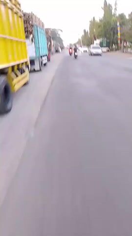 The video shows a view from a moving vehicle on a road. Several large trucks are parked on the left side of the road. In the distance, there are other vehicles, including cars and motorcycles, traveling in the same direction. The environment appears to be a roadside with trees and some buildings. The sky is overcast. The camera is angled downwards and slightly to the left, capturing the road surface and the side of the parked trucks. There is a license plate visible at the bottom of the frame.