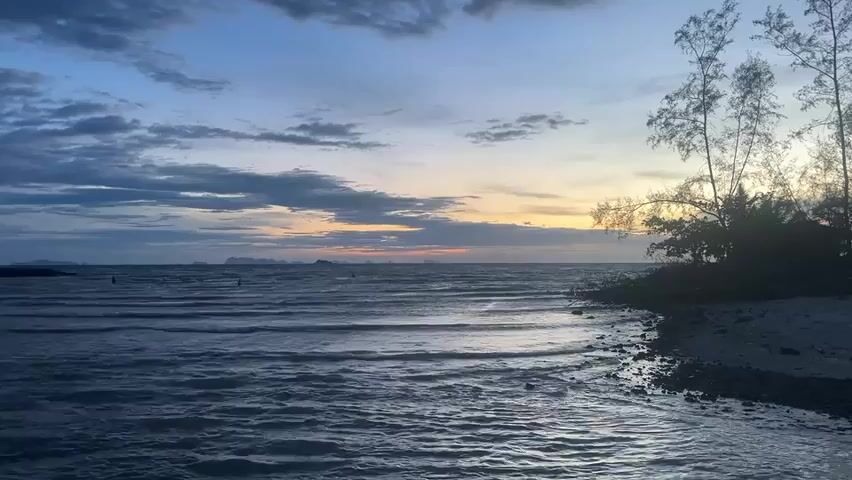 A static shot of a beach and ocean under a cloudy sky at dusk. The ocean is calm, with small waves rolling towards the shore. In the distance, the horizon is visible with a faint orange glow from the setting sun.  To the right of the frame, there is a small beach with dark rocks and light-colored sand, partially obscured by silhouetted trees and vegetation. A few small buoys are visible in the water.