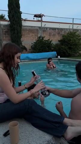 The video shows a woman sitting on the edge of a swimming pool, holding a bottle of liquor and drinking from it. Two men are in the pool behind her, and another person is partially visible in the water. There is a wall and some trees in the background. The scene appears to be an outdoor gathering.