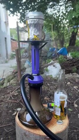 The video shows a hookah smoking on a wooden stump outdoors. The hookah is purple and black, with a foil-covered bowl on top and a black hose. Smoke is billowing from the hookah. Next to the hookah is an open bottle of Corona beer, a lighter, and the bottle cap. The background includes a building, trees, and various debris scattered on the ground. The scene appears to be in a backyard or outdoor area.