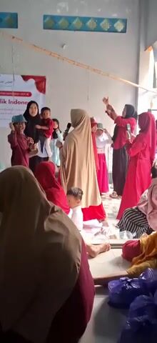 The video shows a group of people, predominantly women and children, gathered indoors. The room appears to be a communal space, with a concrete floor and simple walls. In the background, a banner is visible with Indonesian text and red and white accents, suggesting a celebratory or national event. People are dressed in traditional Islamic attire, including hijabs and long dresses. Some are holding what appear to be small objects, and one woman is holding a microphone. Children are present and seem to be participating in the activities. The overall atmosphere appears to be festive and communal.