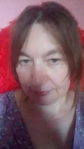 A woman with shoulder-length brown hair and blue eyes, wearing a purple floral-patterned top, looks directly into the camera. A fluffy red object is visible behind her head, and the background is a soft pink. Her expression is neutral, with a slight smile.