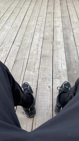 The video shows a first-person perspective of someone sitting on a wooden deck. Their black pants and black sneakers are visible. The camera is pointed downwards, focusing on the person's legs and feet, and the weathered wooden planks of the deck. The scene is outdoors, and the lighting suggests daytime. There are no other people or objects clearly visible in the frame, apart from the person's attire and the deck. The person's legs move slightly, and their feet are positioned apart.
