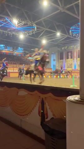 A performance is taking place inside an arena with spectators seated in tiered bleachers. The arena floor is covered in sand. Several horse riders, dressed in colorful costumes, are participating in the event. One rider in the foreground, wearing a blue and yellow outfit with a red sash, is on a dark-colored horse and is raising their arms. Another rider, also in a similar costume, is on a lighter-colored horse in the background. There are flags and poles set up on the arena floor. Decorative lights are suspended from the ceiling, including circular light fixtures with strings of lights. The foreground features a wooden barrier decorated with yellow drapes. The overall atmosphere suggests a circus or historical reenactment performance.