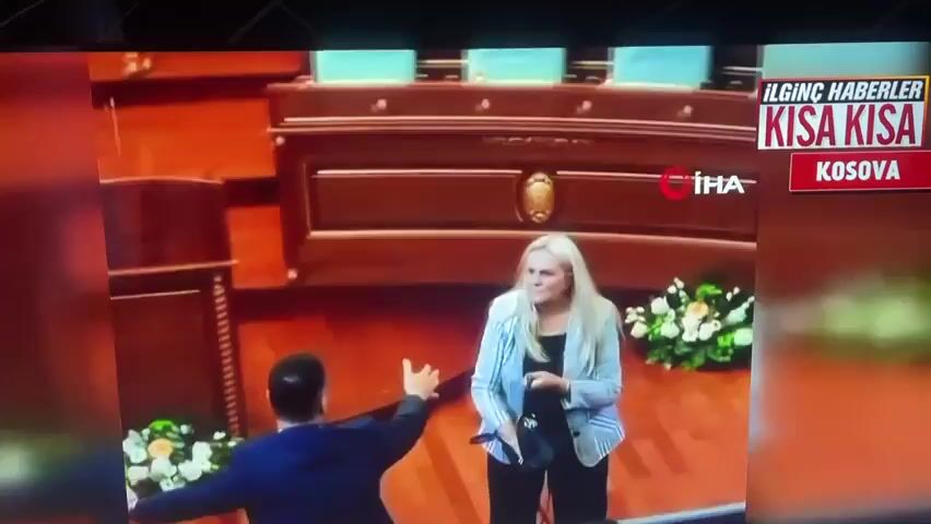 A woman with blonde hair is in a formal setting, possibly a parliamentary chamber, with wooden desks and chairs visible in the background. She is wearing a light blue blazer over a black outfit. A man in a dark blue suit is reaching out and touching her arm, seemingly trying to restrain her. Another man in a suit is visible in the foreground, partially obscured. The woman appears to be agitated, with her arm raised. There are floral arrangements to the right of the frame. A news graphic in the upper right corner reads "ILGINC HABERLER KISA KISA KOSOVA", indicating the report is from Kosovo and features brief, interesting news. The IHA logo is also visible.