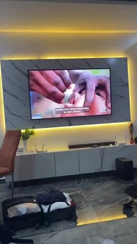 The video shows an indoor scene, likely a modern living room, with a large flat-screen television mounted on a wall. The wall features a grey marble-like pattern and is accentuated by yellow LED strip lighting around the TV and beneath a white TV console.

On the TV, a video is playing. At the beginning, it displays a close-up of hands administering eye drops to a child. Shortly after, the scene changes to show a group of people, including adults and children, gathered outdoors in what appears to be a rural setting. A red basket and a bicycle are visible. Overlayed on this scene is the text "FOR EVERY CHILD A HEALTHY LIFE", and the UNICEF logo is seen in the top right corner, suggesting the content is a public service announcement or documentary related to children's welfare.

Below the TV, a white console holds various items, including a soundbar. To the left, a tall, orange-brown upholstered chair and a small round table are visible. The floor is tiled with a grey and white patterned marble. In the foreground, an open black suitcase with clothes spilling out of it sits on the floor, along with a black bag next to it, suggesting packing or unpacking activities. A black subwoofer is positioned on the floor to the right of the console. The room is brightly lit by artificial light.