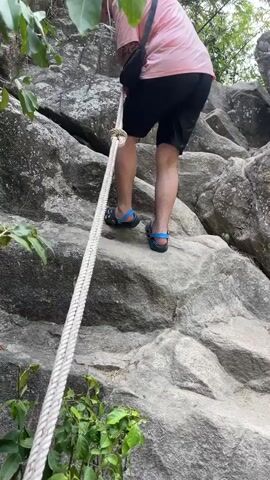 A person is climbing up a rocky incline. The individual is wearing a pink shirt, black shorts, and black sandals with blue accents. They have a black bag slung across their body. A thick, light-colored rope is anchored above and held by the person to assist in the climb. The rock surface is grey and uneven, with some patches of green moss or vegetation. Trees and other greenery surround the rock face. The camera angle looks up at the person from a slightly lower position.
