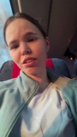 A woman sits in a bus seat, facing the camera. She has fair skin, light brown hair pulled back, and wears a white T-shirt and a light blue fleece jacket. The bus seat is patterned in blue and red. There's a window to her left showing the outside, although details are unclear. She appears to be talking or singing along to music, as her lips are moving. The lighting is somewhat dim.