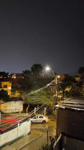 The scene is an urban street at night, under heavy rain. A streetlight illuminates the immediate area, highlighting the downpour. Electrical wires crisscross in the foreground, reflecting the light. A white car is parked on the wet street. Buildings of varying heights are visible in the background, some with lights on. Vegetation, including trees and palms, is interspersed among the buildings. The overall scene is dark and wet, typical of a nighttime rainstorm.