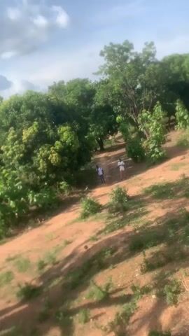 The video shows two people standing on a dirt path in a rural area. The path winds through trees and low-lying vegetation. The ground is mostly reddish-brown dirt with patches of green grass. The sky is partly cloudy, with a blue sky peeking through the clouds. The people appear to be conversing, but their words are not audible.