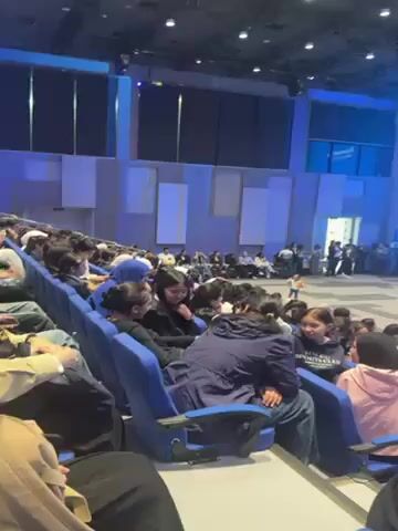 The video shows a large auditorium filled with people seated in rows of blue chairs. The room is brightly lit with ceiling lights. At the front of the auditorium, there is a large screen displaying text and images. A few individuals are visible on the stage in front of the screen. The audience members are looking towards the stage, and some are holding up their phones, possibly recording or taking pictures. The atmosphere appears to be that of a presentation or a conference. There is music playing in the background.