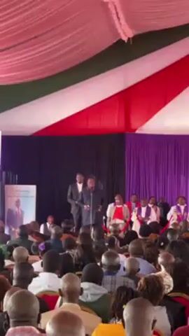 The video shows a man addressing a large audience from a podium under a tent decorated with red, white, and green fabric. Behind him, seated on a platform, are several people in religious vestments. A banner to the left of the speaker features a portrait of another man. The audience, mostly composed of African individuals, is listening attentively. The setting appears to be a formal gathering, possibly a religious or community event.