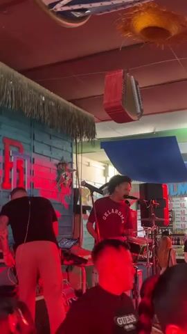 The video captures a live music performance inside a lively bar or restaurant in Fuengirola, Spain, likely during the evening or at night. On a brightly lit stage with blue wooden paneling and green/red accent lighting, two musicians are performing. One musician, wearing a burgundy t-shirt with "OKLAHOMA" printed on it, is actively playing a drum set and singing into a microphone. To his left, another musician in a black t-shirt is operating a keyboard or DJ equipment.

Hanging above the stage are various decorative elements, including a straw-like hat and a distinctive red box-shaped object. In the background, a bar area is visible, stocked with bottles and featuring multiple signs that prominently display "5.0". Audience members are visible in the foreground, with some individuals' backs illuminated by green and blue stage lights. The scene suggests a vibrant and engaging entertainment venue.