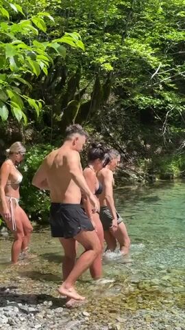 Four people are standing in shallow, clear blue water near a rocky shore. Two women and two men are present. The woman on the far left wears a white bikini and sunglasses. The woman in the center wears a black bikini. One man in the center wears black swim shorts and appears to be flexing his arms. Another man is to his right, also in swim shorts. The background is a lush green forest with trees and foliage. The sun is shining, creating bright light and shadows. The water is calm, with small ripples visible. The scene appears to be a natural outdoor setting, likely a river or lake.