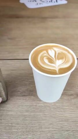 A hand sprinkles cinnamon from a glass shaker onto a paper cup of latte with latte art on a wooden surface.