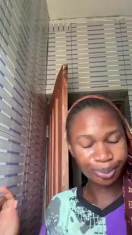 A young woman is standing in a room. She is facing the camera and has a maroon headscarf partially covering her hair. She wears a teal and purple patterned shirt. She is making faces at the camera and sticking out her tongue. The wall behind her is covered in a horizontally-striped light gray and blue tile pattern. To her right is a dark-colored door that is partially open, revealing a dark space behind it.