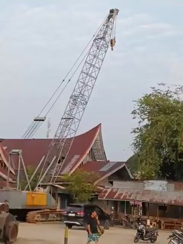 The video opens with a view of a large construction crane with its boom extended high into the sky. In the background, there are several buildings with red, angled roofs. A person is walking in the foreground. The scene then shifts to a white building with red and white trim and a flagpole adorned with red and white banners. Next, the video shows a group of people standing around, with some individuals appearing to be engaged in conversation. A tall tower, possibly another crane or structure, is visible in the distance. The final segment of the video shows a group of people gathered on a dock or pier, with a body of water and some buildings in the background. Several motorcycles are parked near the buildings. The overall environment appears to be an outdoor area, possibly a construction site or a port, with a mix of structures and people.