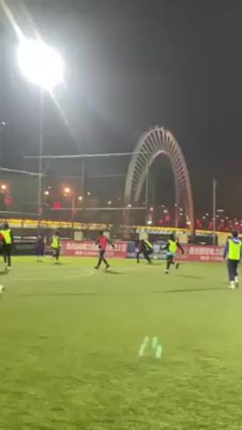 The video shows a soccer game being played at night on a green turf field. A large, white arch bridge is visible in the background, illuminated by lights. The field is lit by bright floodlights. Several players wearing yellow vests and red jerseys are moving around the field, kicking a soccer ball. One player falls to the ground. A few white water bottles are visible in the foreground. There is fencing surrounding the field, and banner advertising is displayed along the sidelines. Traffic and buildings are seen in the background behind the fence.
