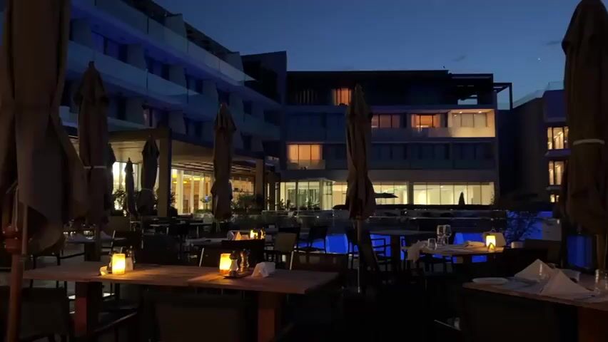 The video shows an outdoor restaurant seating area at dusk. Several tables are set with white tablecloths and lit by candles. Umbrellas are lowered over some of the tables. In the background, a modern building with illuminated windows is visible, and a pool of water with blue lighting. The sky is a deep blue with hints of pink and orange from the setting sun. The overall atmosphere is calm and serene.