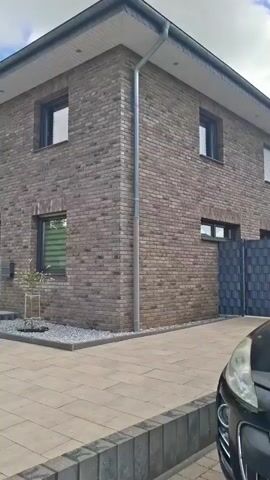 The video pans across the exterior of a modern brick house. The house has a dark brick facade and a white overhang. There are several windows with dark frames. A gray downspout runs vertically along the corner of the house. The front door is glass with an arched top. A small tree is planted in a gravel bed near the entrance. The ground in front of the house is paved with large rectangular stones. To the left, a red brick walkway leads towards a street with cars and trucks. The sky is blue with scattered white clouds.
