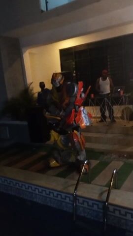 The video depicts a lively party scene at night, centered around a pool and a building. In the foreground stands a person in a detailed Transformer Bumblebee costume, striking a pose. To the right, a DJ in a white tank top and a red bandana is operating a laptop on a folding table, likely providing music for the gathering. Other individuals can be seen milling about near the entrance of the building, some dancing and interacting with each other. One person is recording with a phone. The setting is well-lit, with a pool edge visible in the bottom of the frame, adding to the party atmosphere. The overall impression is that of a fun, outdoor celebration.