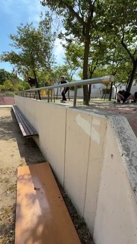 A skateboarder attempts a trick on a ledge but falls. The skateboarder is wearing a black t-shirt and black pants. The skateboard is green and yellow. The skateboarder is in a park with trees, benches, and concrete structures. Another person is visible in the background sitting on the ground. The sky is blue with some clouds.