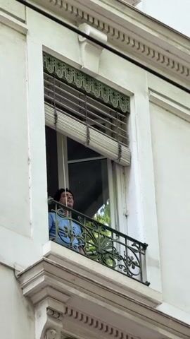 A man is captured in a medium shot, standing on a small balcony of a white, classically styled building, located in Lyon, France. He has dark hair and is wearing a light blue long-sleeved shirt. The balcony features an ornate black metal railing and is situated in front of a window with a white frame and a partially rolled-up beige Venetian blind.

The man is seen looking outwards, and a faint cloud of vapor or smoke is visible near his mouth. He then looks down, holding and interacting with a smartphone. Throughout the video, he appears relaxed and content, subtly moving his head to the rhythm of music playing in the background. The audible music sounds like Latin American or Spanish popular music, with the phrase "mi patria querida" being discernible. The scene is brightly lit, indicating it is daytime. No visible text is present on the building or objects within the frame.