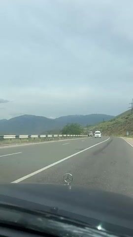The video captures a first-person perspective from a moving vehicle driving along a highway through a mountainous landscape. In the immediate foreground, the dark hood of the car is visible, featuring a prominent Mercedes-Benz emblem. The asphalt road ahead has clear white lane markings.

At the beginning of the video, a white van is seen approaching from the opposite direction on the left lane, passing the camera's vehicle. Beyond the road, a silver guardrail runs along the left side, separating the highway from a gentle slope with green vegetation. The background is dominated by a range of hills and mountains, suggesting an elevated or rural setting. The sky is overcast, filled with clouds, indicating a cloudy daytime with diffused light. No other specific people are visible, and no additional text is legible besides the car's emblem. The scene evokes a sense of travel through a scenic, natural environment, consistent with the provided location context of Vardashat, Armenia.
