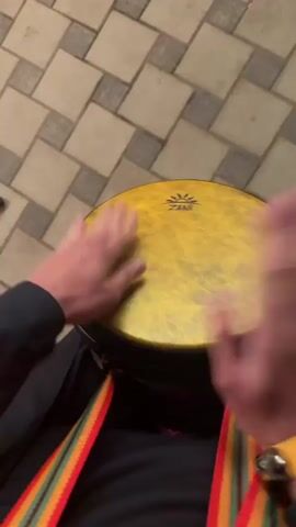 A close-up, overhead shot shows a person playing a djembe drum. The drum is yellow with a black rim and a visible "ZANI" logo. The person's hands are blurred from the rapid motion as they strike the drumhead. A colorful, striped strap, featuring red, yellow, and green, is visible around the person's neck or chest. The person is also wearing a yellow wristband with several small bells attached. The background consists of a patterned tile floor, with some red lighting reflected on the tiles. The shot is framed from the perspective of someone looking down at the drum and hands.