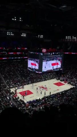 The video shows an overhead shot of a basketball game in an arena. The court is brightly lit and surrounded by fans. A large screen hangs above the center of the court, displaying the game footage. Players in red and blue uniforms are actively playing. The crowd is audible.