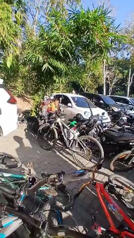 The video shows a group of people gathered around bicycles and cars outdoors, possibly in a parking lot or staging area. The setting appears to be sunny with trees in the background. Several mountain bikes are visible, some leaning against each other and others parked upright. One person in the foreground, wearing a blue and white cycling jersey and a black helmet, is interacting with another person who has a yellow helmet. They seem to be engaged in a conversation or activity related to their bikes or the event. Other individuals in similar cycling attire are also present in the background, along with a line of cars. The overall impression is of a cycling event or group preparing for an activity.